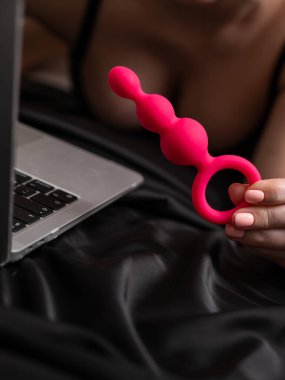 Laptopun yanında pembe anal boncuklar tutan kadın siyah çarşafın üzerinde yatıyordu.