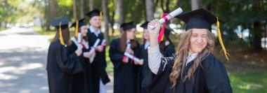 Mezuniyet elbisesi giymiş bir grup mutlu öğrenci dışarıda. Ön planda elinde diploması olan genç bir kız.