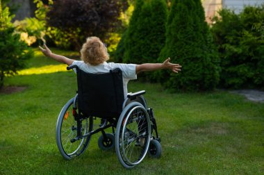 Rear view of an elderly woman spread her arms to the sides while sitting in a wheelchair on a walk outdoors