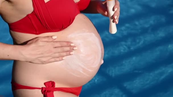 孕妇穿着红色比基尼晒日光浴时的腹部特写 准妈妈涂防晒霜 — 图库视频影像