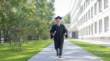 Mezuniyet elbisesi giymiş yaşlı bir adam dışarıda yürüyor ve diplomasını tutuyor.