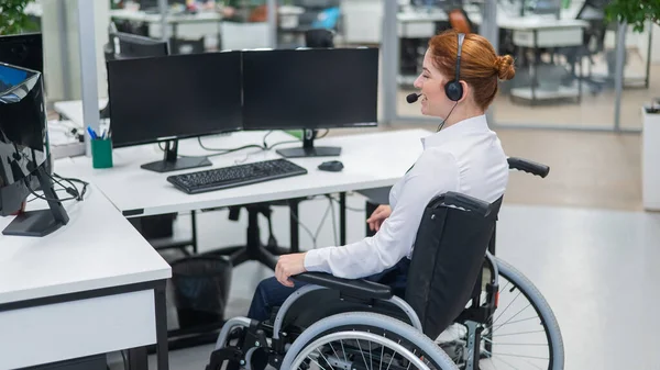Red-haired caucasian woman in a wheelchair talking on a headset. Female call center worker at her desk