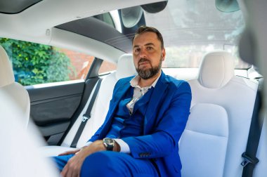 A caucasian man in a blue suit sits in the back seat of a car. Business class passenger