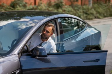 Beyaz sakallı, takım elbiseli bir adam yazın kırsalda siyah bir elektro arabadan iniyor.