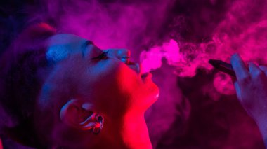 Tıraşlı şakakları olan Asyalı bir kadın neon ışıkta sigara içiyor.