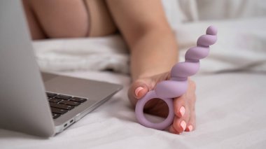Yatakta yatarken dizüstü bilgisayarın yanında leylak anal boncuklar tutan kadın.