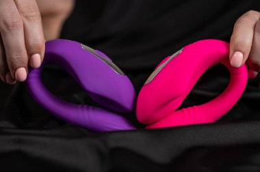 Bir kadın siyah ipek çarşafın üzerinde yatarken pembe ve mor eğimli bir seks oyuncağı tutuyor.