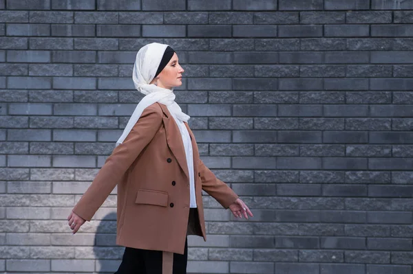 ヒジャーブを着てビジネスススーツを着た若い女性がレンガの壁に沿って歩きます — ストック写真