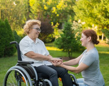 Beyaz bir kadın, tekerlekli sandalyede oturan yaşlı bir annenin elini tutuyor. Dışarı çık.