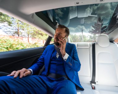 A caucasian man in a blue suit is talking on the phone in the back seat of a car. Business class passenger