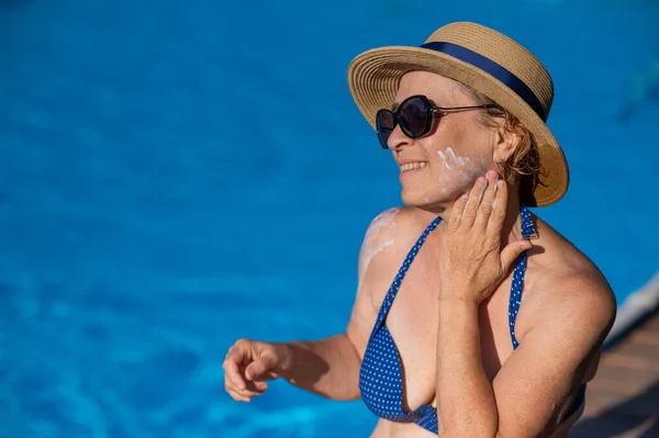 Portrait of an old woman in a straw hat, sunglasses and a swimsuit applying sunscreen to her face while relaxing by the pool