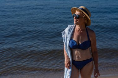 Hasır şapkalı, güneş gözlüklü ve mayosu olan yaşlı bir kadın sahilde dinleniyor.