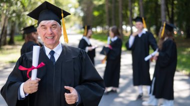 Bir grup mezun açık havada bornozla geziyor. Yaşlı bir öğrenci diploma aldığına sevinir.