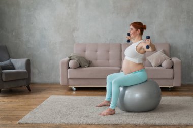 Hamile kadın evde spor yaparken dambıllarla egzersiz yapıyor.