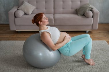Hamile kızıl saçlı kadın evde fitball ile kol egzersizi yapıyor.