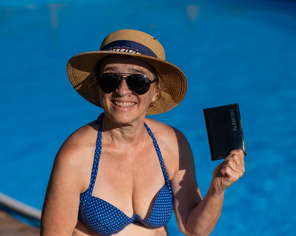 An elderly woman in a hat and sunglasses holds a passport while sitting by the pool. Vacation in retirement