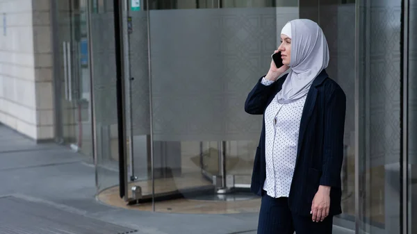 Pregnant business woman in hijab and suit talking on smartphone while leaving business center