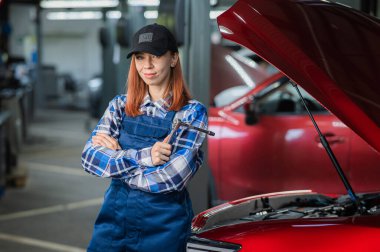 Kadın bir oto tamircisi arabanın açık kaputunda duruyor ve elinde bir tornavida tutuyor.