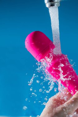 Bir kadın mavi arka planda akan suyun altında pembe bir vibratörü yıkıyor. Seks oyuncağı hijyen konsepti
