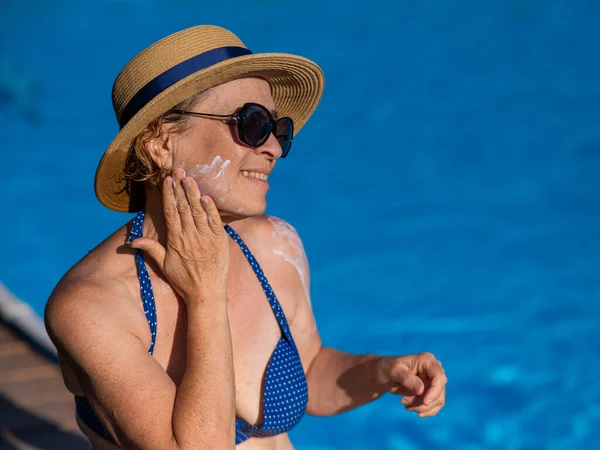 Portrait of an old woman in a straw hat, sunglasses and a swimsuit applying sunscreen to her face while relaxing by the pool