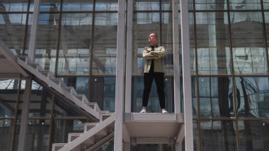 Haki gömlekli bir adam kollarını göğsüne bağlamış bir şekilde bir gökdelenin önünde duruyor.