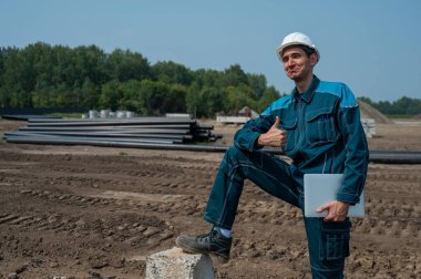 İnşaat işçisi dizüstü bilgisayar tutuyor ve inşaat alanında dikilirken baş parmağını gösteriyor.