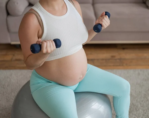 孕妇坐在健身球上 一边用哑铃做运动 怀孕的腹部的特写 — 图库照片