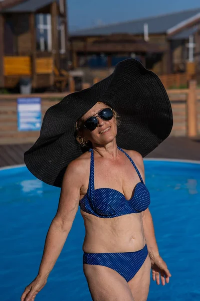 An elderly woman in a huge straw hat and sunglasses is sunbathing by the pool. Retiree on vacation