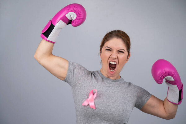 A woman with a pink ribbon on her chest raises her hands in pink boxing gloves on a gray background. Victory over breast cancer