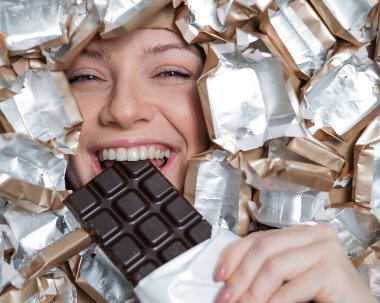 The face of a Caucasian woman surrounded by candy wrappers. The girl eats a bar of chocolate