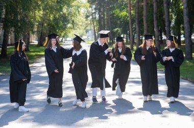 Mezuniyet elbisesi giymiş mutlu gençler. Öğrenciler parkta yürüyor.