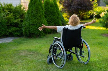 Rear view of an elderly woman spread her arms to the sides while sitting in a wheelchair on a walk outdoors