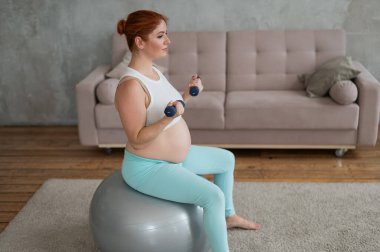 Hamile kadın evde spor yaparken dambıllarla egzersiz yapıyor.