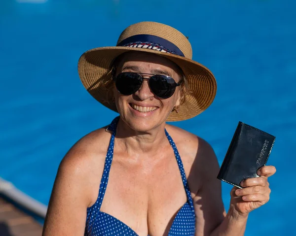 An elderly woman in a hat and sunglasses holds a passport while sitting by the pool. Vacation in retirement