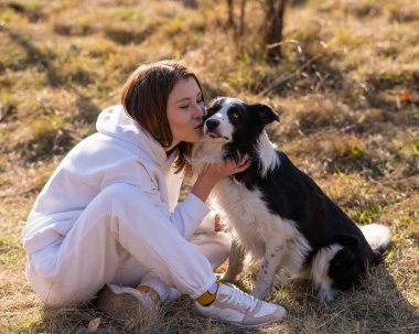 Beyaz bir kadın sonbahar parkında çimenlerde otururken Collie köpeğine sarılıp öpüyor.