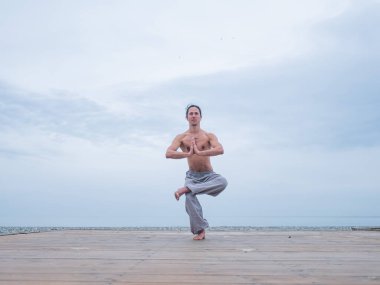 Beyaz bir adam, çıplak gövdesiyle deniz kıyısında wushu yapıyor.