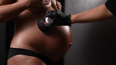 Usta, bronzlaştırma işleminden sonra hamile bir kadının cildine ışıltıyla pudra sürer. Karnına yakın çekim.