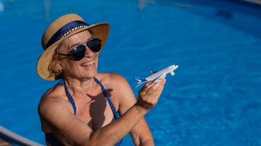 Şapkalı ve güneş gözlüklü yaşlı bir kadın havuz kenarında otururken model bir uçak tutuyor. Emeklilik tatili kavramı