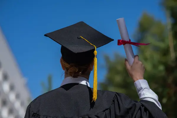 Stock image Rear view of a caucasian woman in a graduate gown holding a bundle with a diploma and standing near the university