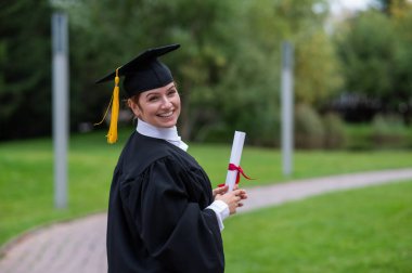 Mezuniyet elbisesi giymiş, açık havada diploma taşıyan mutlu beyaz kadın portresi.