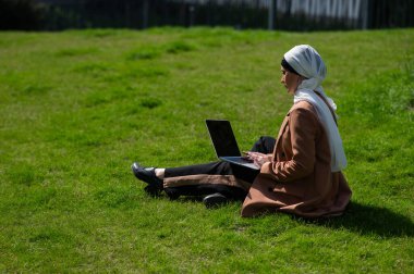Başörtüsü takan genç bir kadın çimlere oturur ve dışarıda dizüstü bilgisayar kullanır.
