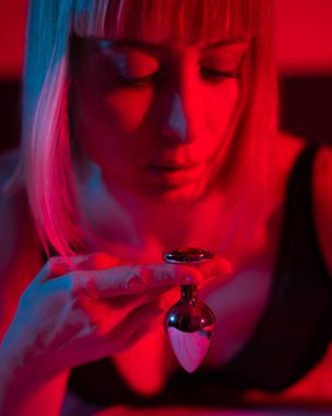 The blonde lies on the bed in red blue neon light and holds a steel butt plug