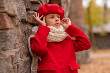 Kırmızı paltolu ve bereli üzgün bir beyaz kız tuğla duvara yaslanmış duruyor.