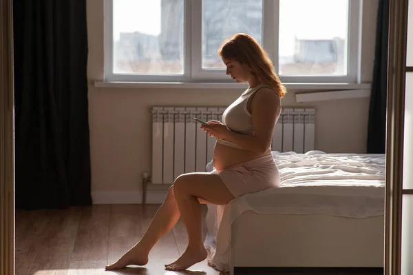 Μια Έγκυος Γυναίκα Χρησιμοποιεί Ένα Smartphone Ενώ Κάθεται Στο Κρεβάτι — Φωτογραφία Αρχείου