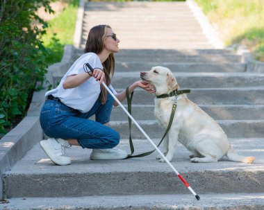 Kör genç sarışın parkta yürürken bir rehber köpeği okşuyor. Merdivenlerde değnekli bir kadın var.