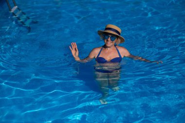 Havuzda mavi gözlüklü ve şapkalı yaşlı bir kadının portresi yüzüyor.