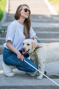 Kör genç sarışın parkta yürürken bir rehber köpeği okşuyor. Merdivenlerde değnekli bir kadın var.