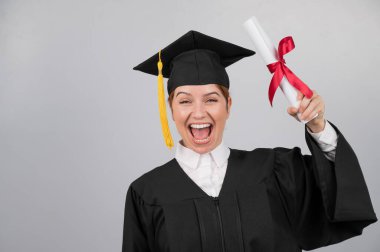 Mezuniyet elbisesi içinde gülümseyen kadın beyaz arka planda diploma taşıyor.