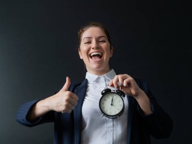 Takım elbiseli mutlu bir bayan öğretmen elinde çalar saatle okul yönetim kurulunda duruyor ve baş parmağını kaldırıyor.