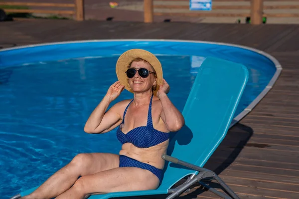 An old woman in a hat and sunglasses is sunbathing on a sun lounger by the pool. Vacation in retirement